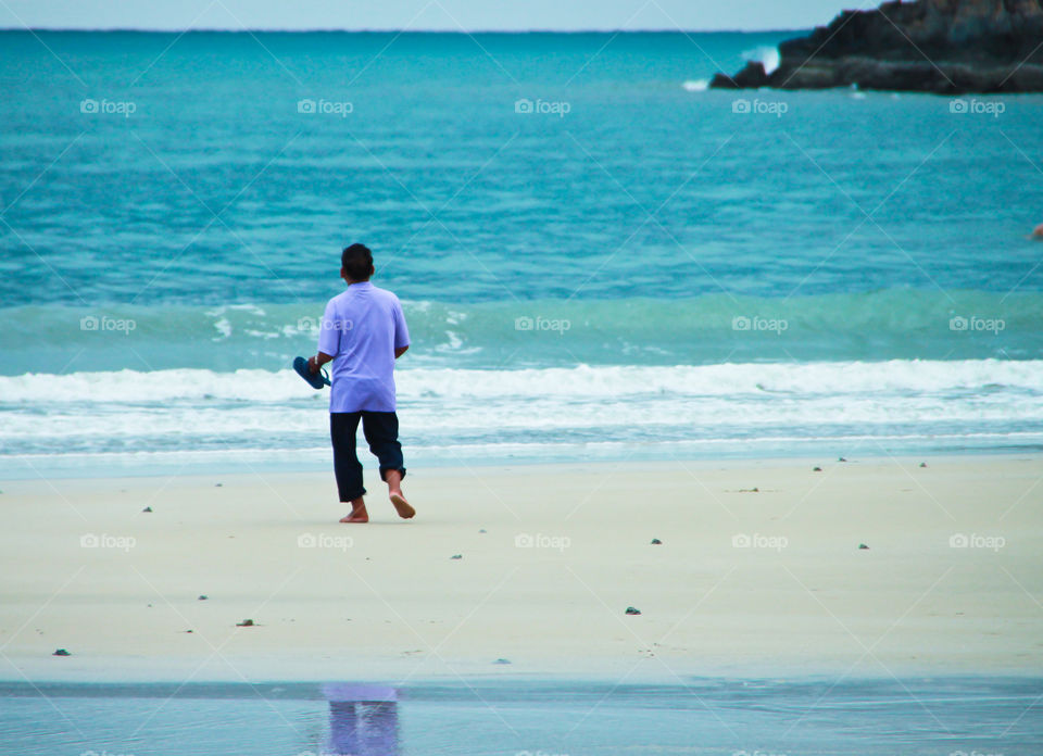 A man walks on the beach holding shoes, walking barefoot on the sand happily.