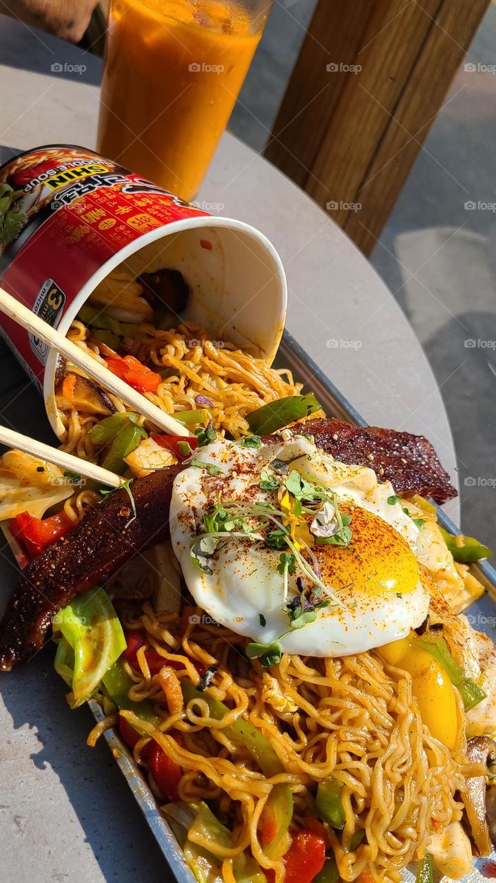 Delicious spicy Asian ramen noodles with sunny side up egg, Thai fusion, cascading out of ramen cup, topped with vegetables and awesome bacon