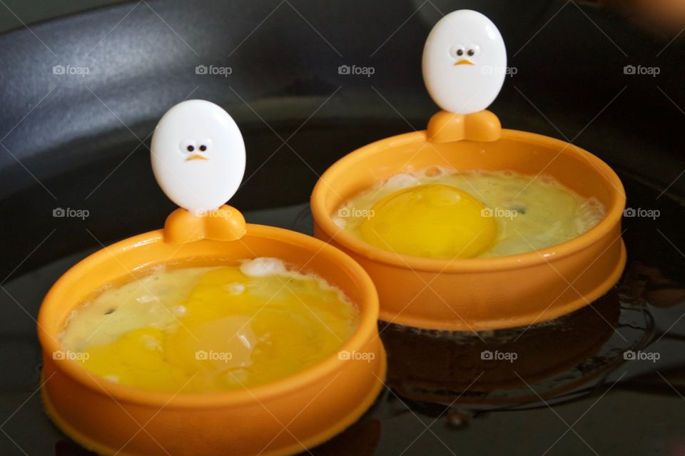 Closeup of a cast iron pan containing eggs being fried in silicone egg rings with egg-shaped handles depicting baby chick faces and feet