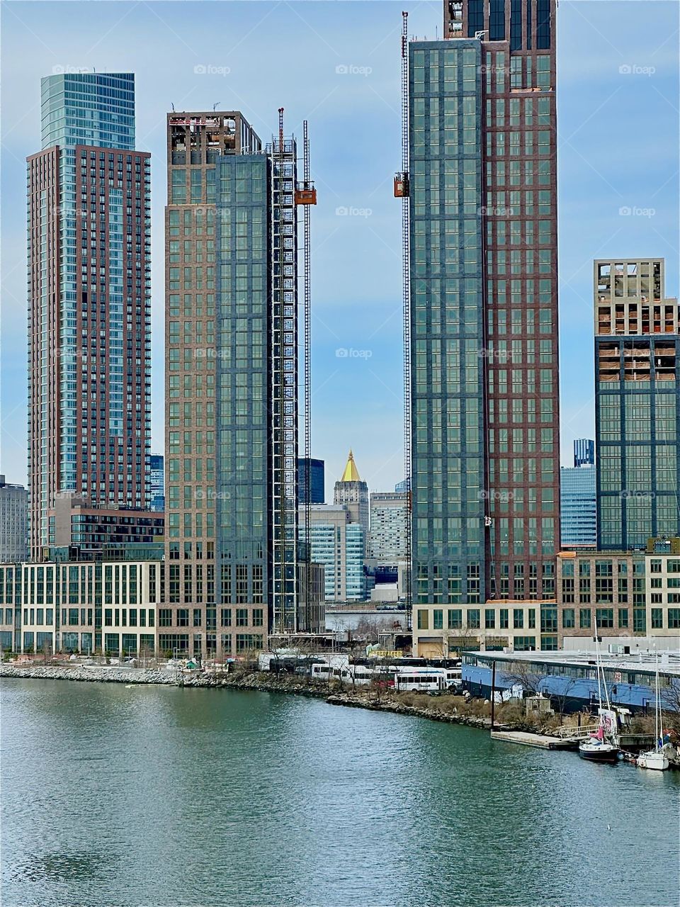 This is „Newtown Creek“ seen from the „Pulaski Bridge“ that connects „Greenpoint“, Bklyn to LIC. Between two recently built high rises with outside elevators we see the „NY Life Bldg“ with the golden roof in „Manhattan“. 2024. Hypnotic Productions