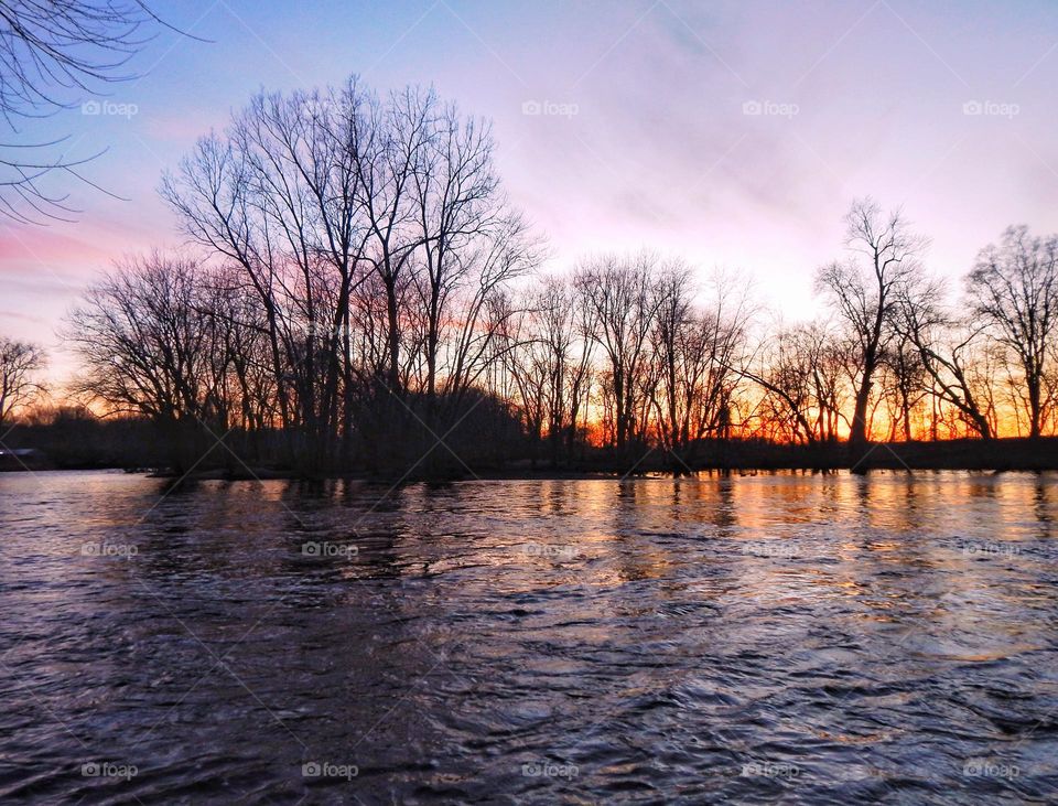 Sun setting on Michigan river turning the sky and clouds shades blue, pink, orange, and purple and silhouetting the trees in a dark shadow against the bright sky. The river shows the bright reflection of the sky and the dark shadows of the trees