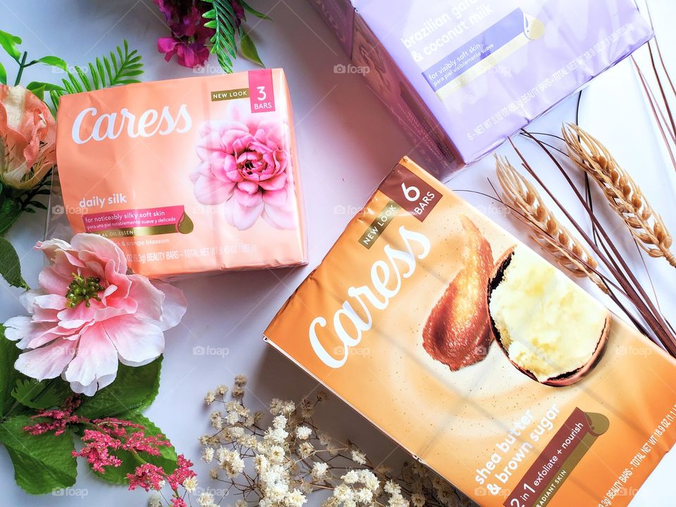 The new look Caress bar including daily silk 3 pack, shea butter and brown sugar six pack and Brazilian gardenia & coconut milk six pack, on a white surface surrounded by matching florals.