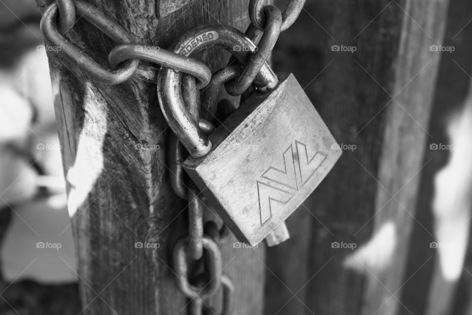 It is an iron chain closed by a padlock. The iron chain is hung on a wooden fence. The photo is taken closely so that the lock and the chain can be seen well.