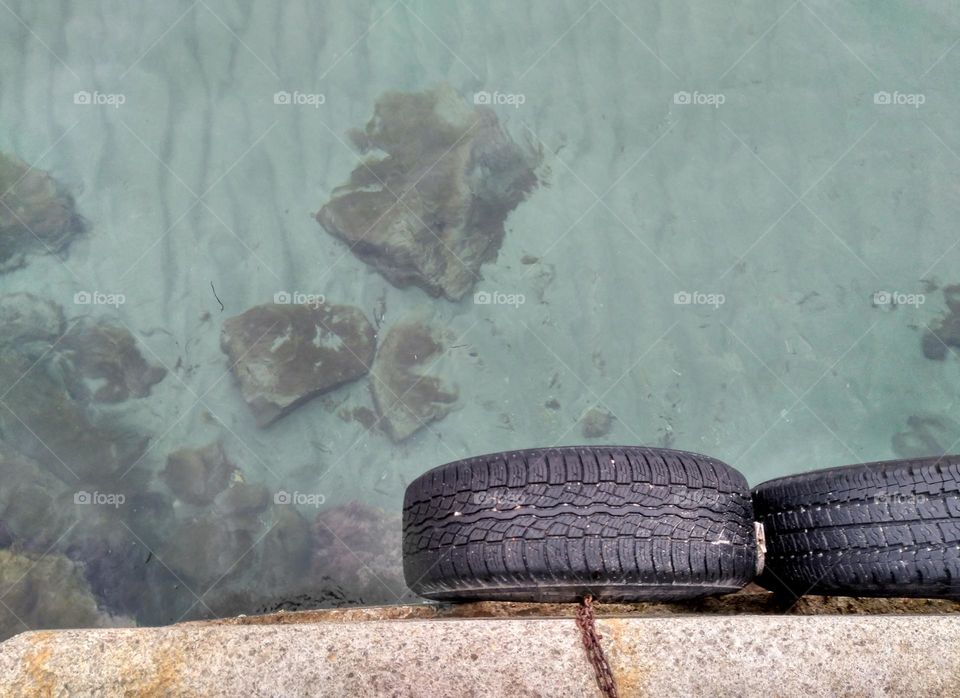 Two tires can be seen tied with rusty chains to a dock in Ibiza, Spain.  In the background the turquoise sea and several rocks within it.