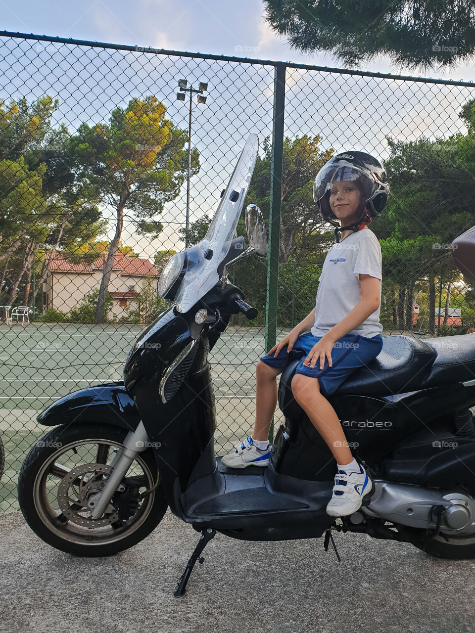 A boy sitting on a motorbike in a helmet on the background of a tennis court.  Summer holidays