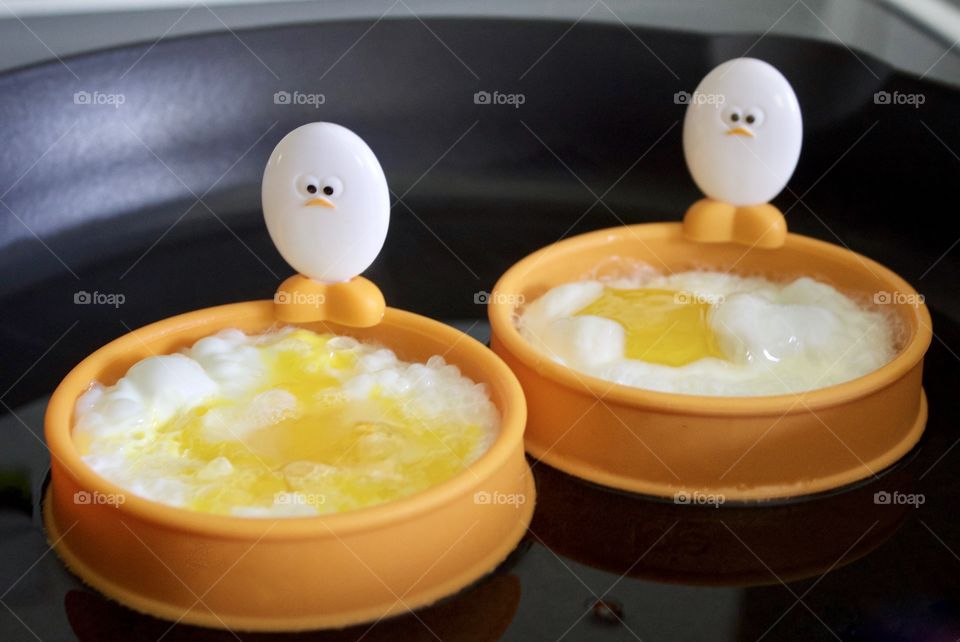 Closeup of a cast iron pan containing eggs being fried in silicone egg rings with egg-shaped handles depicting baby chick faces and feet