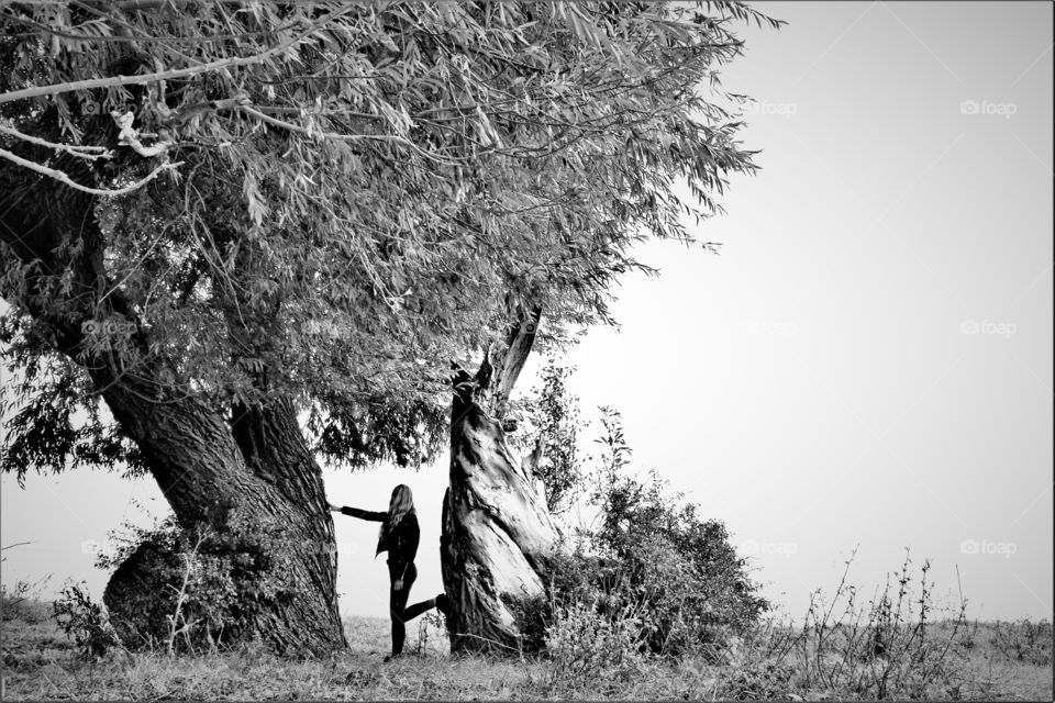 Girl standing between 2 beautiful old trees leaning with her hand and foot against the trunk