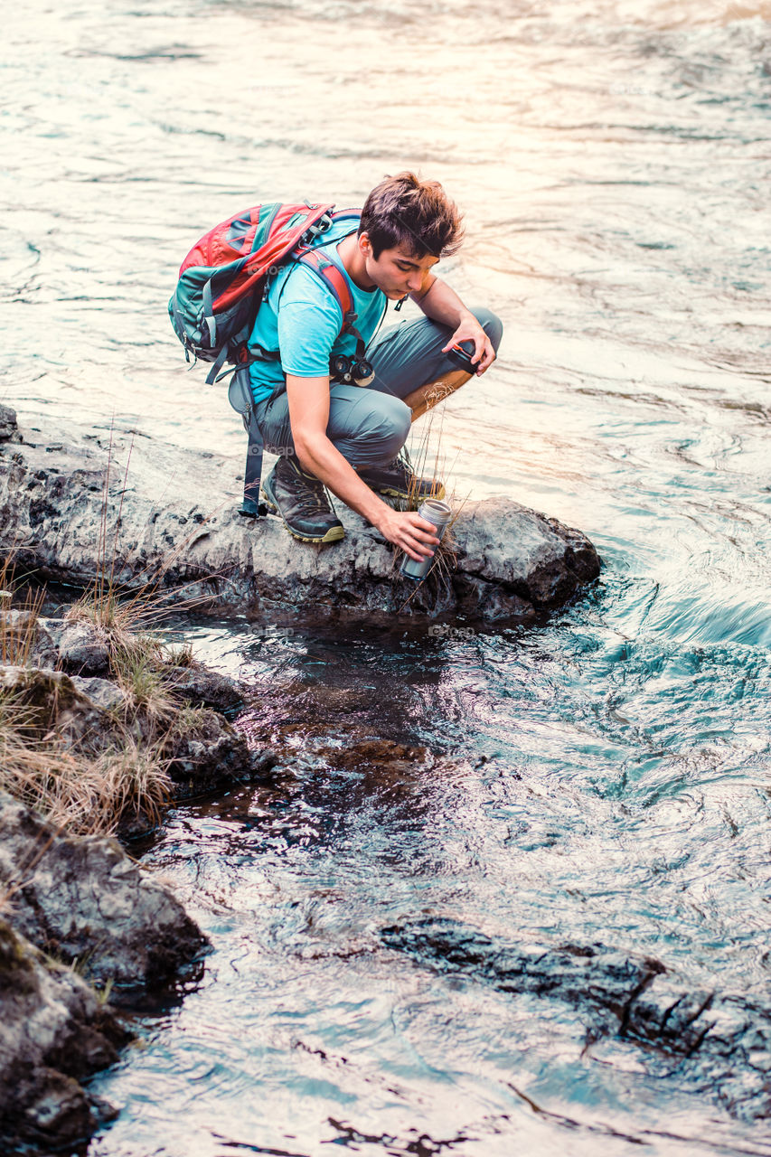 Young wanderer takes pure water from a river into mug. He is sitting on a rock over the river, rests during a hike, spends a vacation on wandering with backpack, he is wearing sport summer clothes