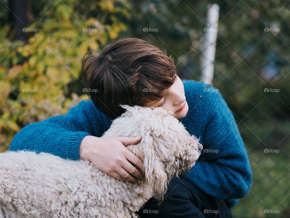 Cute little boy with dark hair wearing blue knitted sweater hugs white curly poodle dog in autumn garden, portrait of child 