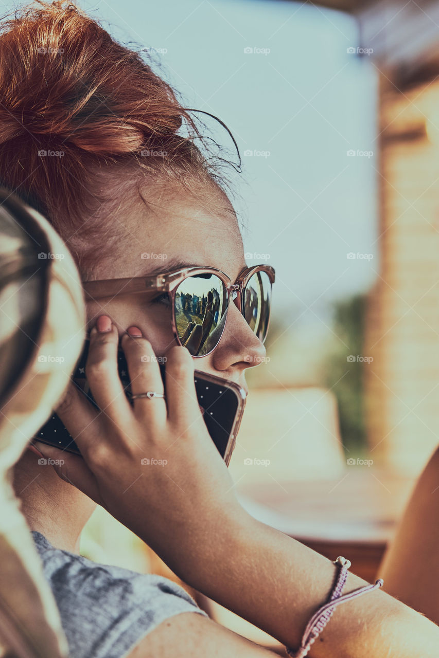 Young woman talking on a mobile phone sitting in a chair outdoors on patio wearing a sunglasses. Real people, authentic situations