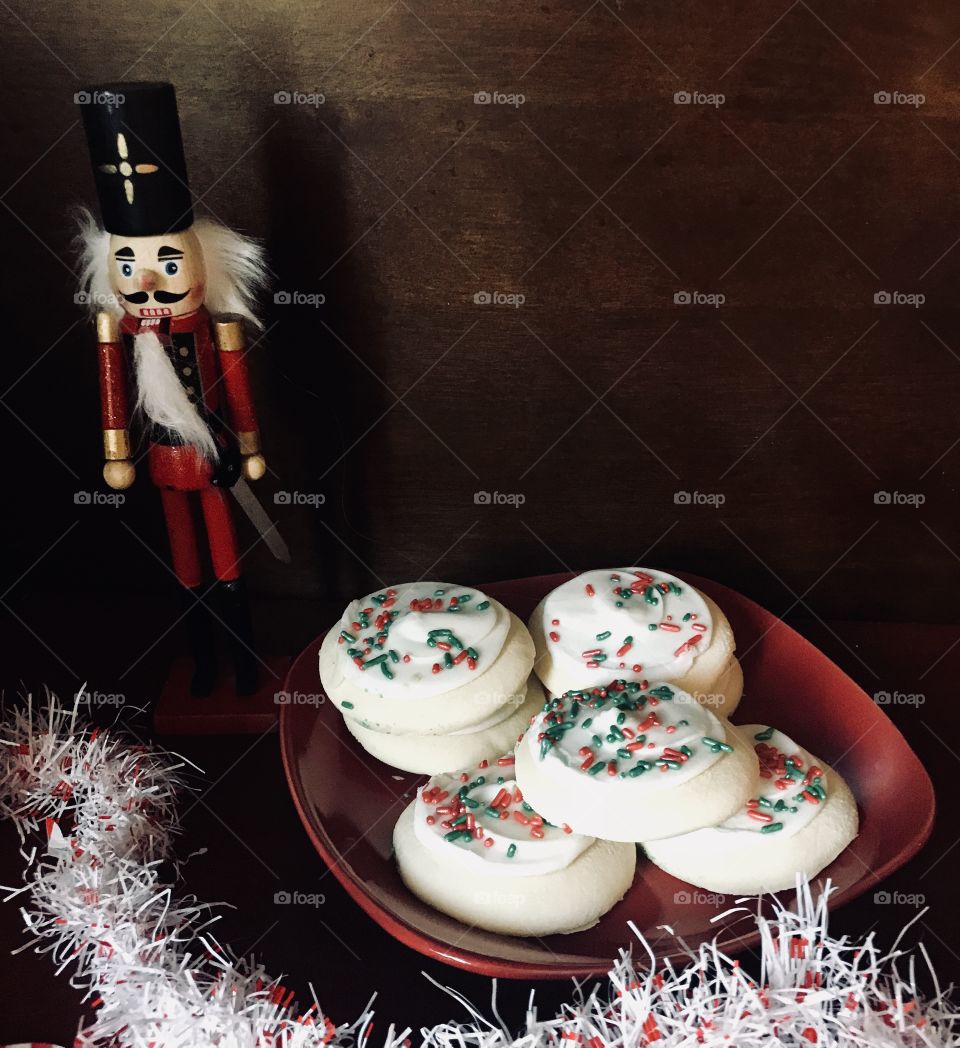 A nutcracker displayed next to a red plate filled with delicious sugar cookies with frosting and colorful red , green and white sprinkles. 