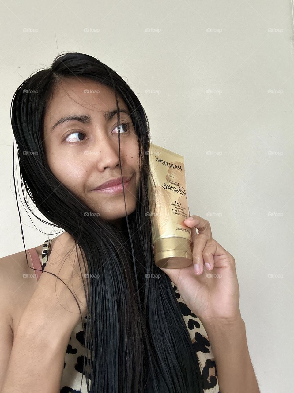 Portrait of a woman with black long hair looking at the left while holding Pantene miracle leave in conditioner. White background. 