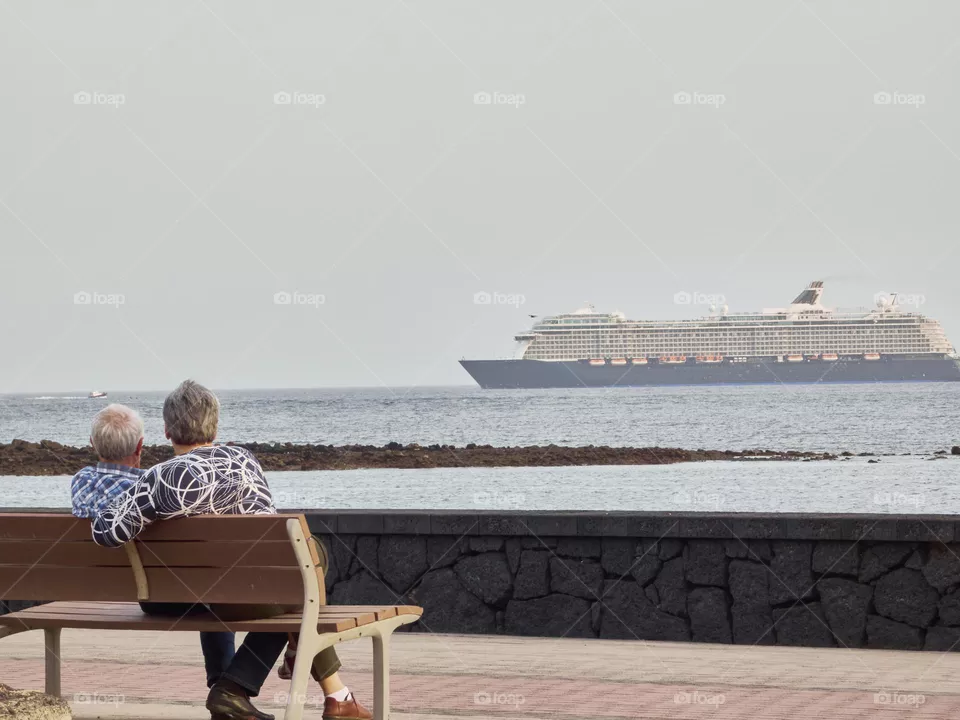pareja de ancianos sentados en un banco con vistas al mar y crucero de fondo