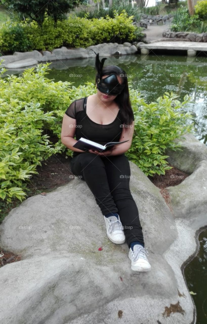 mujer con antifaz o máscara negra sentada con un libro en las manos leyendo, sentada en una roca en un parque