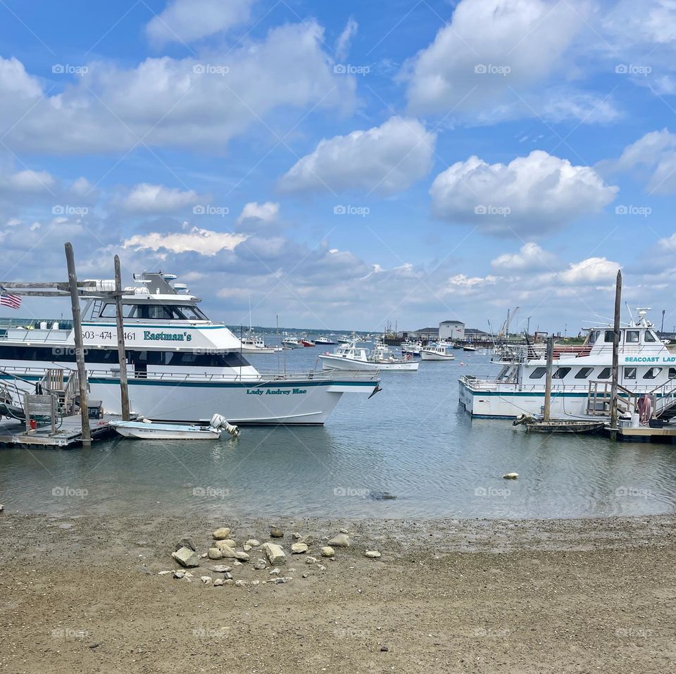A hot July afternoon in Seabrook, NH, with a scenic luncheon view of sailboats and fishing vessels gently bobbing in the sunlit water of Hampton Harbor, framed by blue skies and big white puffy clouds.