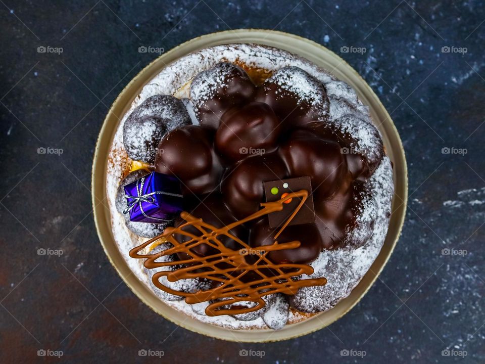 One chocolate cake of profiteroles sprinkled with powder and with a mini gift box in a plate lie in the center on a dark gray stone table, flat lay close-up. Holiday food concept, cakes.