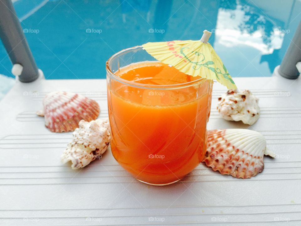 Glass of orange juice with yellow umbrella and seashells near the blue pool water