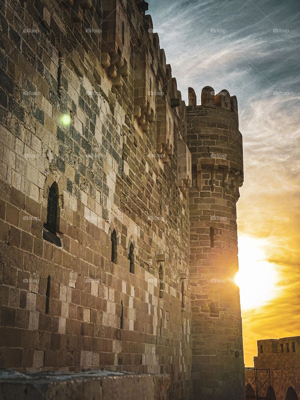 The Qaitbay Citadel in Alexandria, Egypt, stands resolute against a sunset sky, with the sun peeking from behind one of its imposing towers. built in the Mamluk style of architecture.