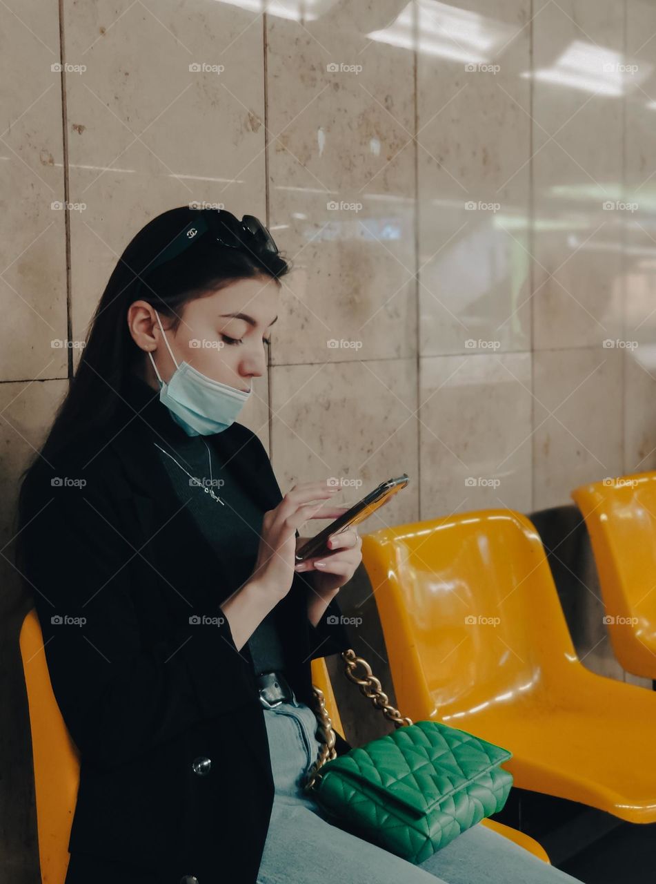 A young beautiful caucasian girl with a medical mask on her face, long hair, in a black jacket, blue jeans, a green handbag on her knees sits on a yellow bench in the subway and holds a mobile phone in her hands flipping the screen with her finger, s