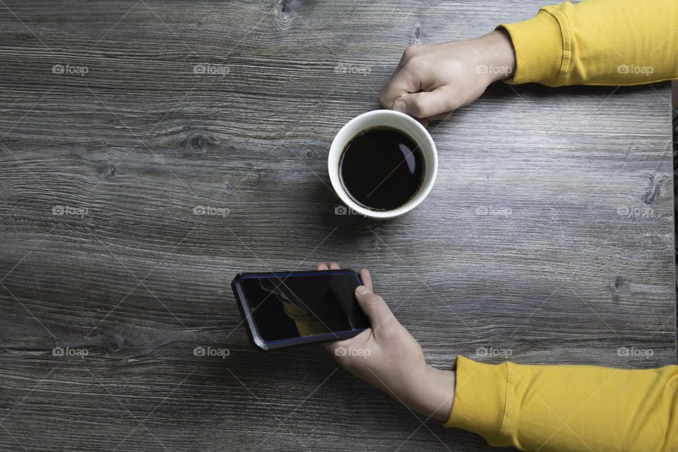 A man drinks coffee in the morning and works on the phone. Men's hands hold a white cup of coffee and a mobile phone in a yellow jacket. The hands lie on a gray wooden surface that is used as a background.