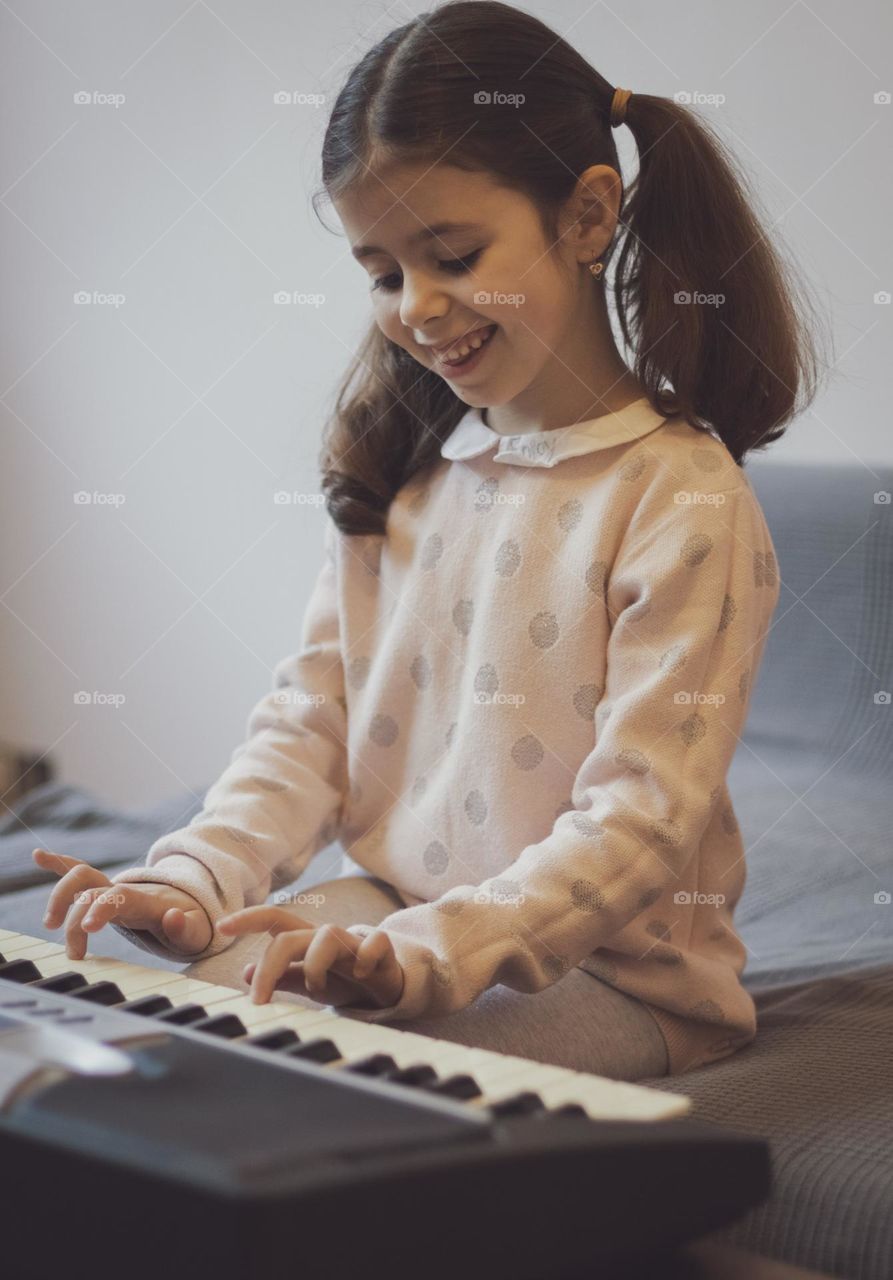 Portrait of a beautiful little caucasian brunette girl with two ponytails on her head with a smile and happy emotions plays the electric piano while sitting on the sofa in the living room, close-up side view. Music education concept, musical instrume