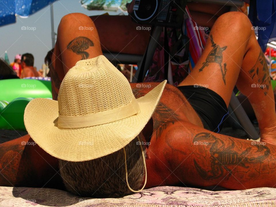 A man is in tattoos, lies on a beach, cowboy shirt, cow-boy, hat, rest