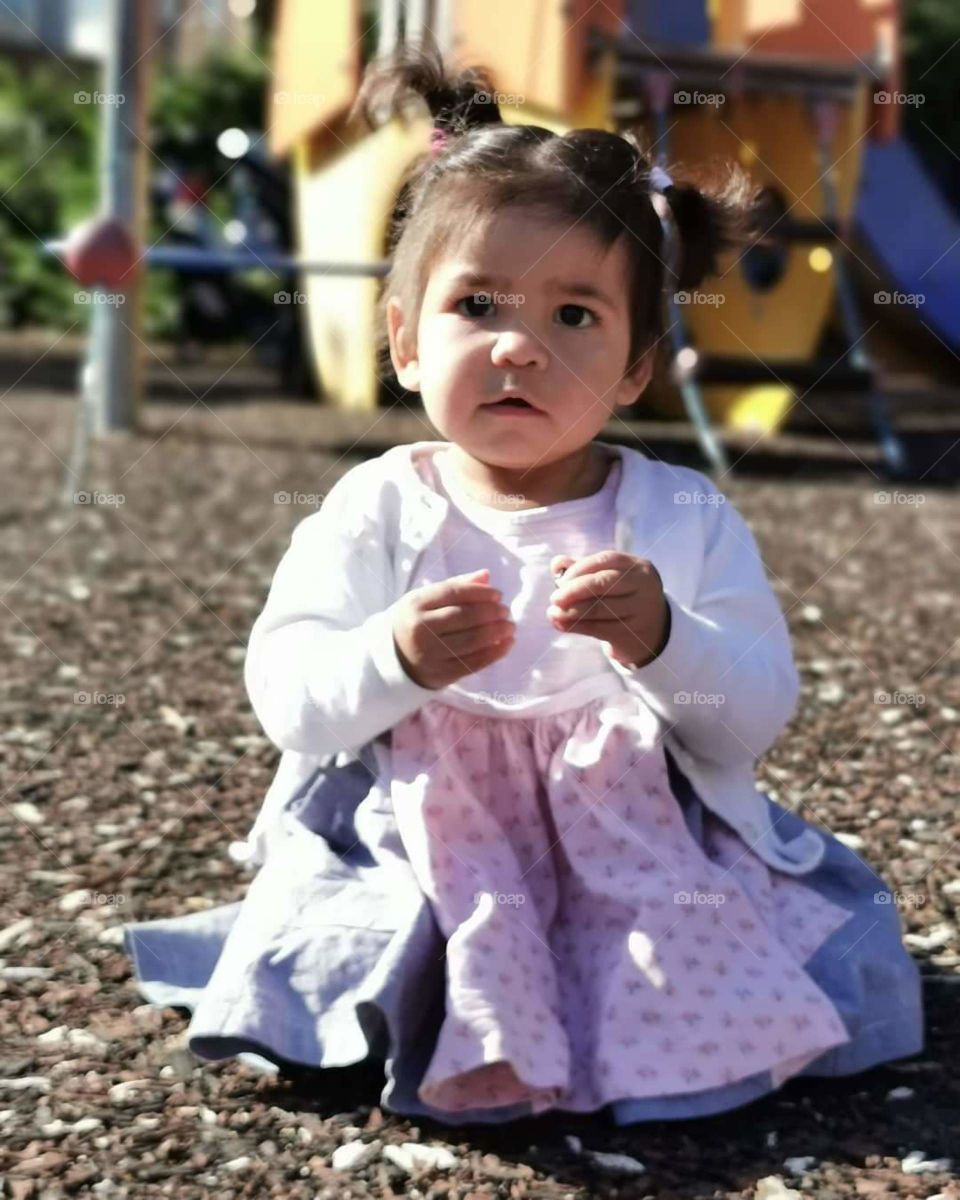 Toddler girl in dress sitting on the ground in the playground. Blurred background.