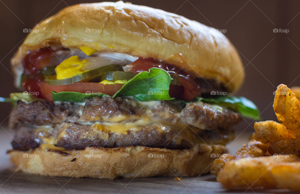 traditional American juicy cheeseburger with lettuce, tomato, onion, pickle, ketchup, and mustard on a sesame bun next to a pile of seasoned fries
