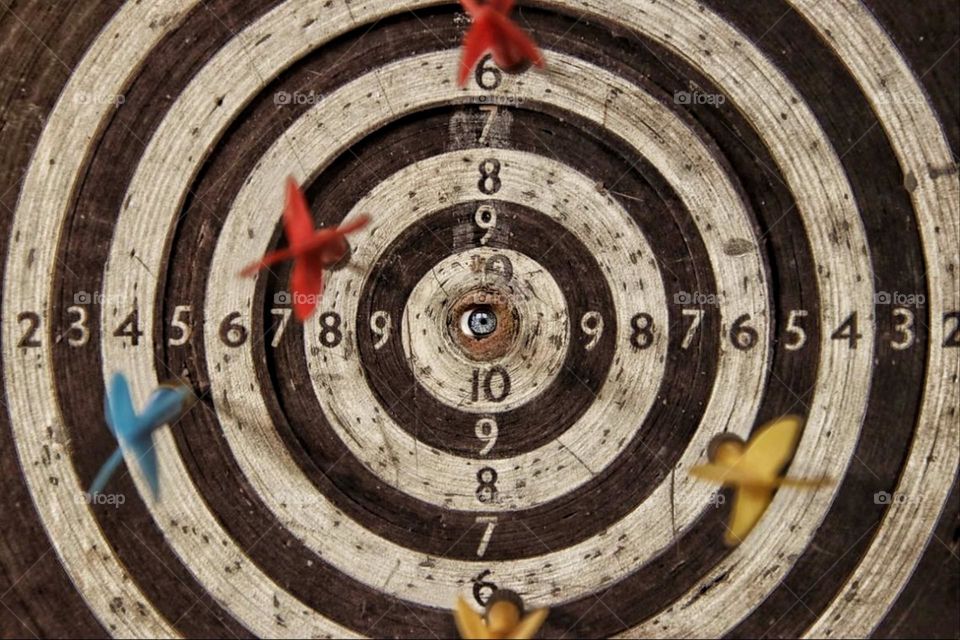 A blue eye looks out of the bulls - eye of an old dartboard framed by yellow and red darts