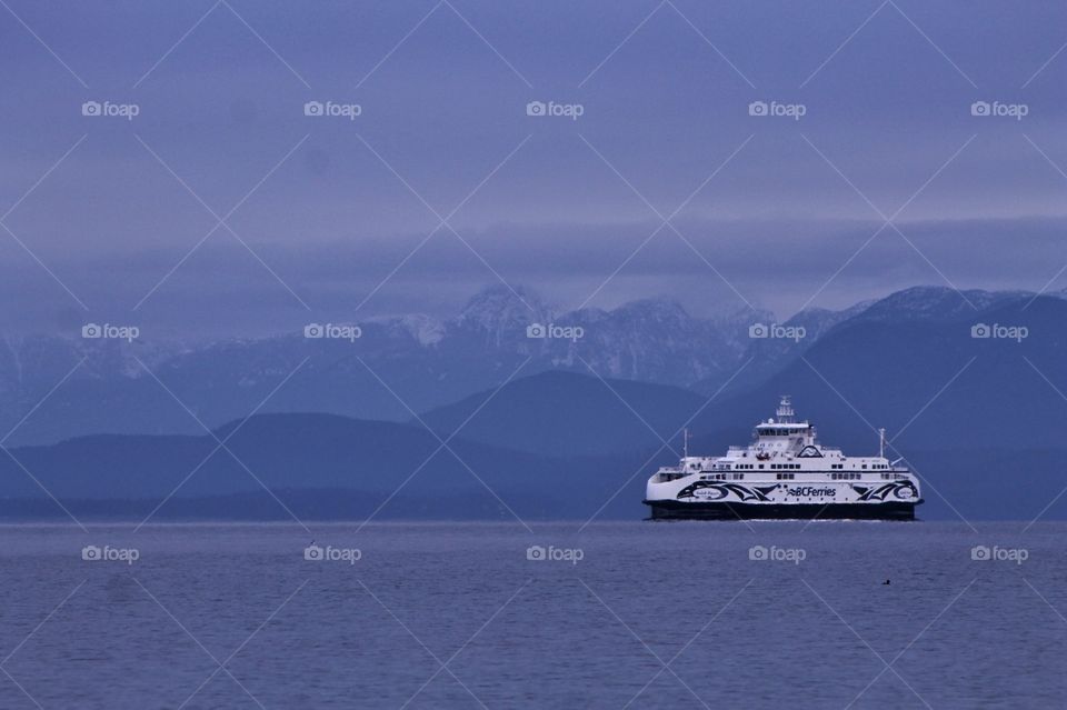 A stark white ferry crossing the ocean strait on calm blue waters. The rolling hills on the coast and the snow capped mountains provide a stunningly beautiful backdrop.