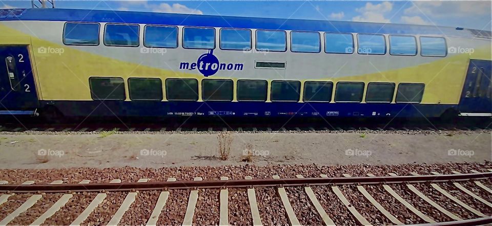 Another view of the train, brightly colored in yellow and indigo blue as well as silvery gray tones from across the tracks. Up closest to us an empty train track can also be seen. 2022. Hypnotic Productions