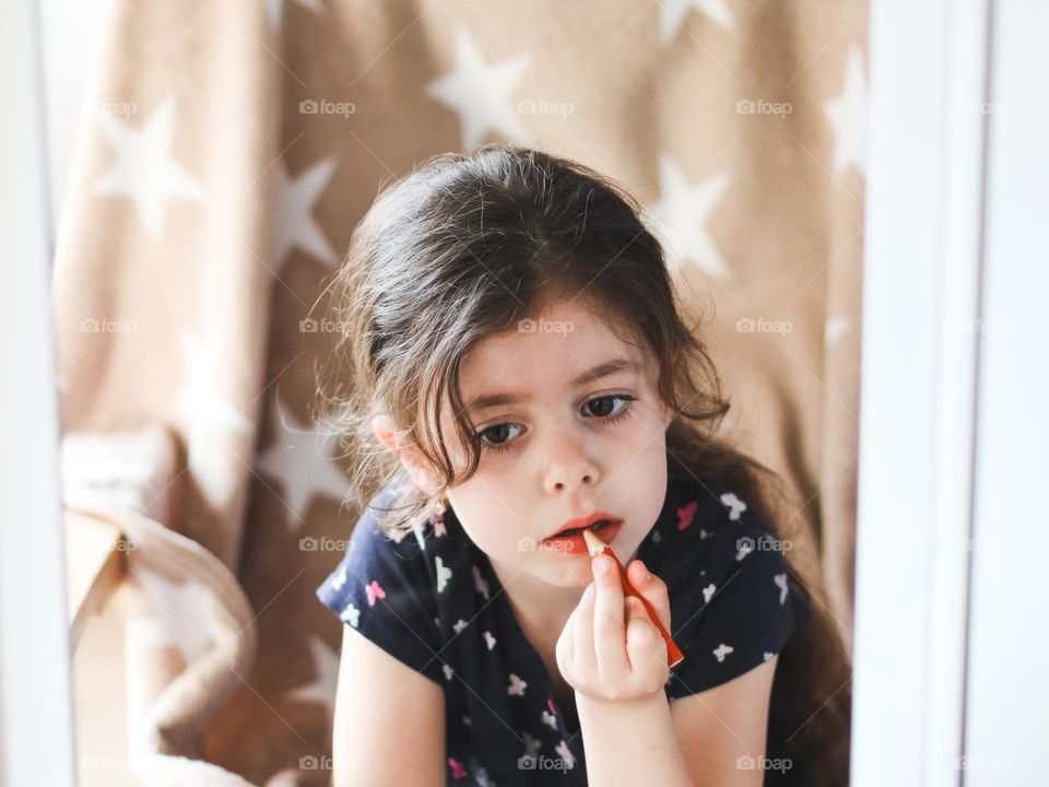 Portrait of a beautiful little caucasian brunette girl painting her lips with a red pencil sitting in a couch with a beige bedspread with stars and is reflected in the dressing room mirror, close-up from the side.