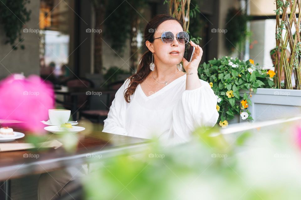 Portrait of smiling beautiful brunette middle aged woman in sunglasses in white clothes with coffee at terrace of the summer cafe