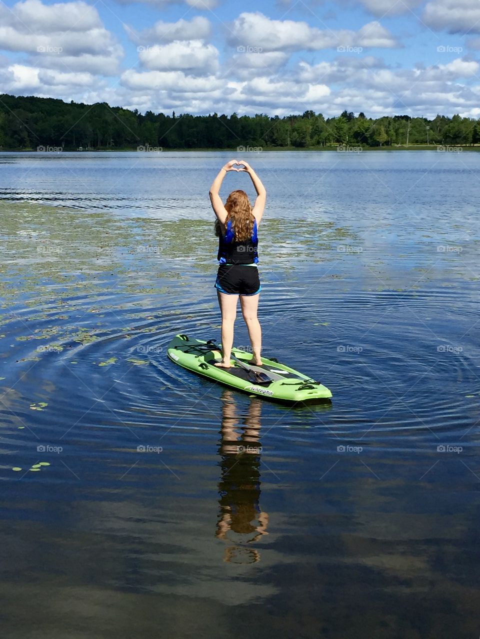 Girl standing on a green paddleboard in a lake with lily pads. She is wearing a blue lifejacket and her hands are up forming a heart above her head. Her reflection is visible in the lake water below.