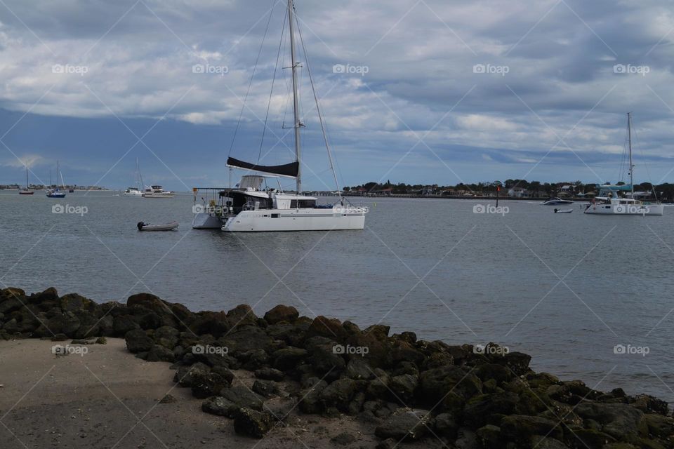 A white sailboat on gray water behind a beach with green vegetation and in front of a blue cloudy sky