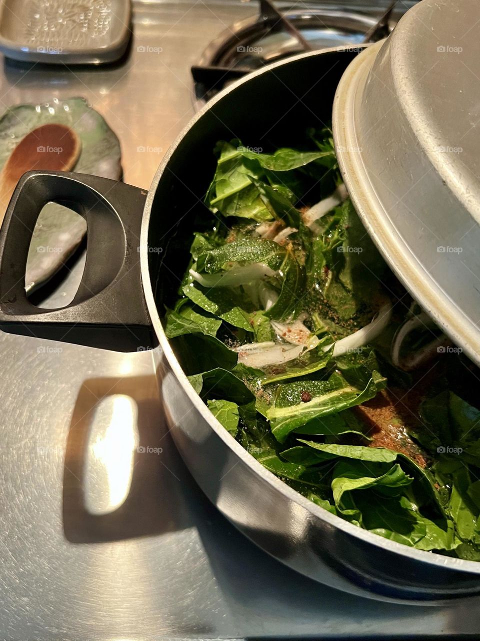 Closeup of chopped collard greens and seasonings in a partially covered pot. Ready to start simmering on a stainless steel stove top.