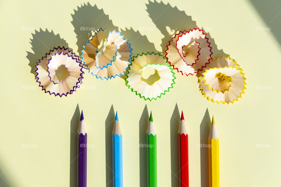 Five 5 colored pencils with wooden shavings are sharpened and lie on a light yellow background with a clear shadow from natural light on a bright, sunny day.