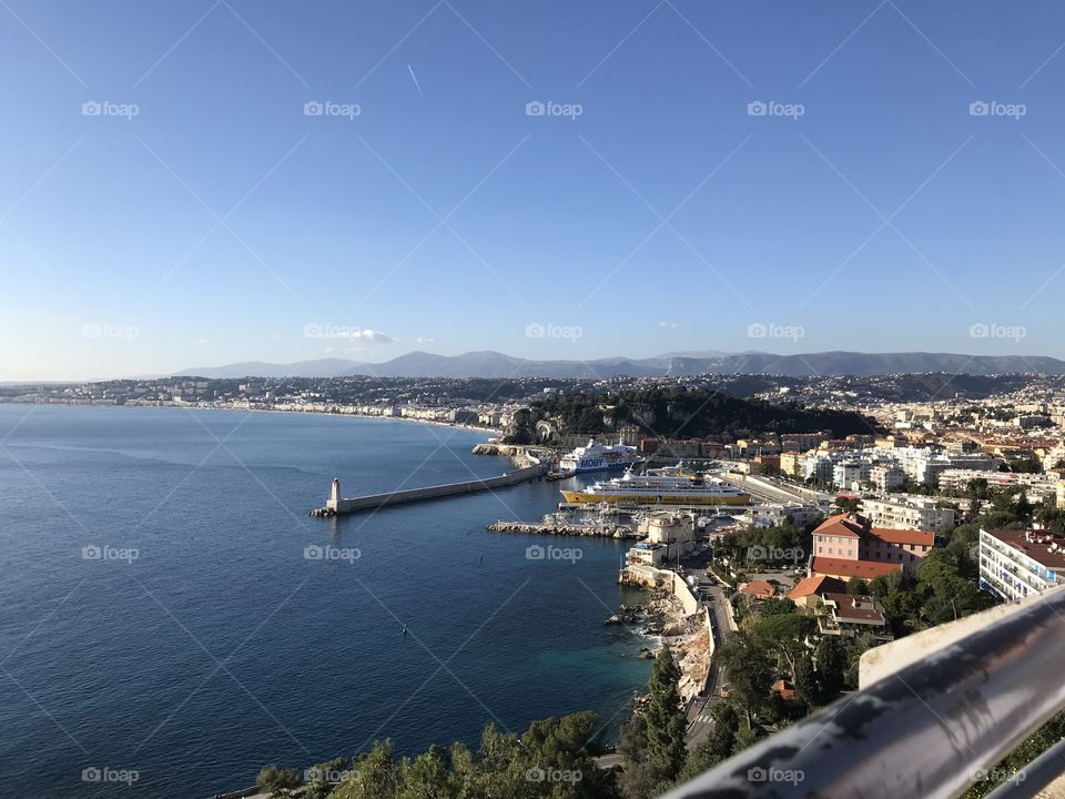 Une magnifique vue de la ville de Nice dans le Sud de la France en janvier 2019. Nous apercevons la mer bleue ainsi que de la verdure sur un fond de maisons. À l'arrière plans, on voit les montagnes.