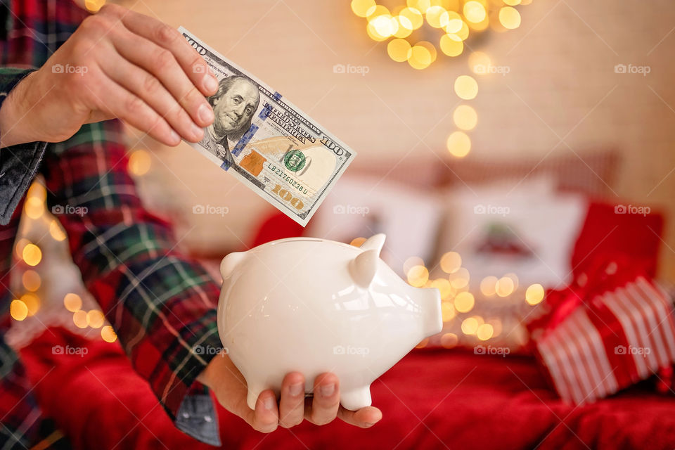 piggy bank with 100 dollars banknote in festive New Year atmosphere of scenery.  Magical bokeh with Christmas tree and bright lights.