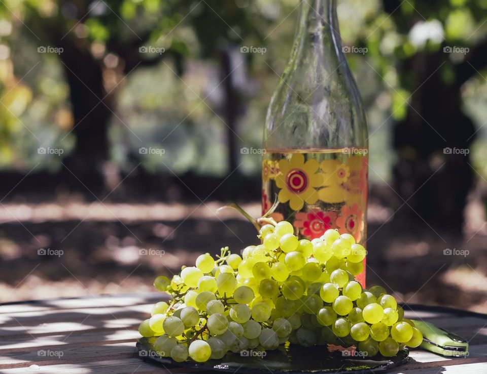 A bunch of grapes with a bottle of wine on a table outdoors in dappled sunlight