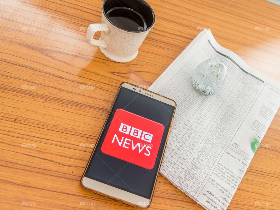 Kolkata, India, February 3, 2019: BBC news app (application) visible on mobile phone screen beautifully placed over a wooden table with a newspaper and a cup of coffee. A Technology Product Shoot.