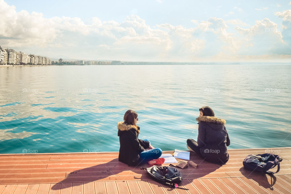 Two Friends Teenager Student Girls Reading Their Books And Talking Outdoor In Front Of The Sea At Port Dock
