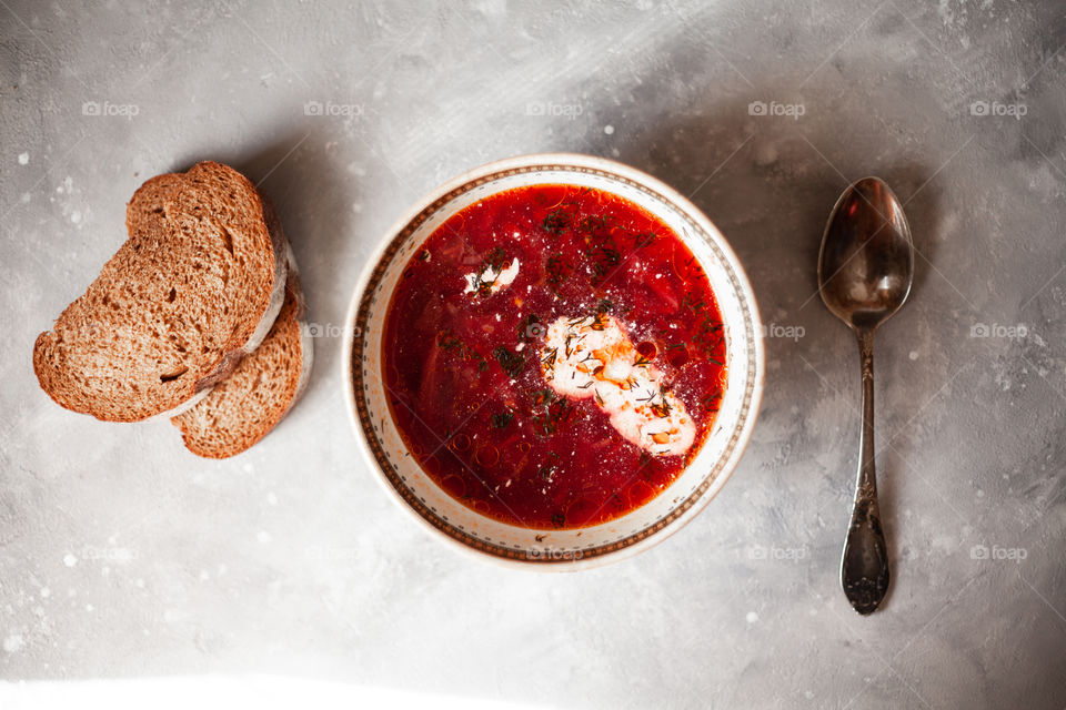 Plate of Ukrainian borsch. Nearby are two sliced ​​pieces of bread and a spoon. A plate is on the newspaper. Photo taken on a white concrete background.Red borsch, add sour cream and greens.