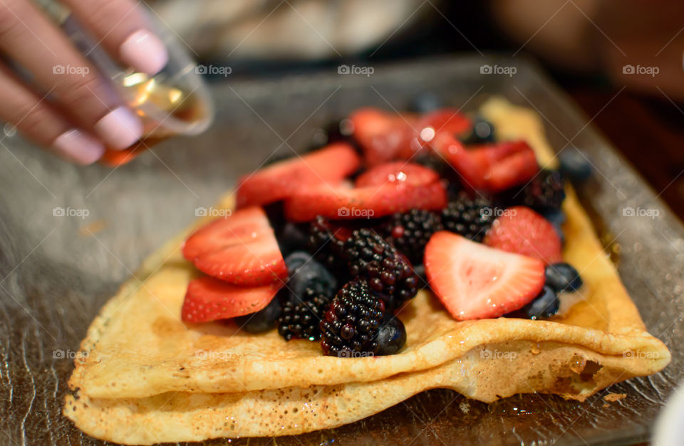Woman pouring maple syrup on a French crepe with a pile of healthy antioxidant rich raw berries including sliced strawberry, blueberry and blackberry fruit topping