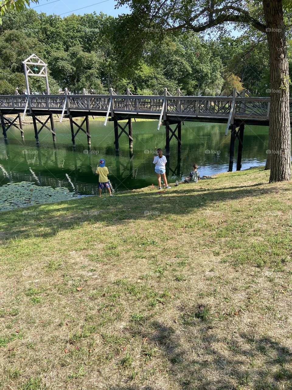 Children fishing in Divine Park in Spring Lake, NJ. Green grass is in the foreground and green trees are in the background. The bridge over the water leads to beautiful walking paths. 