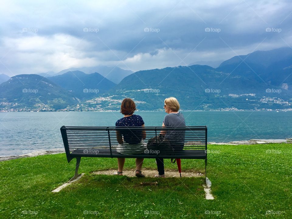 two people sitting on a bench look at Lake Como