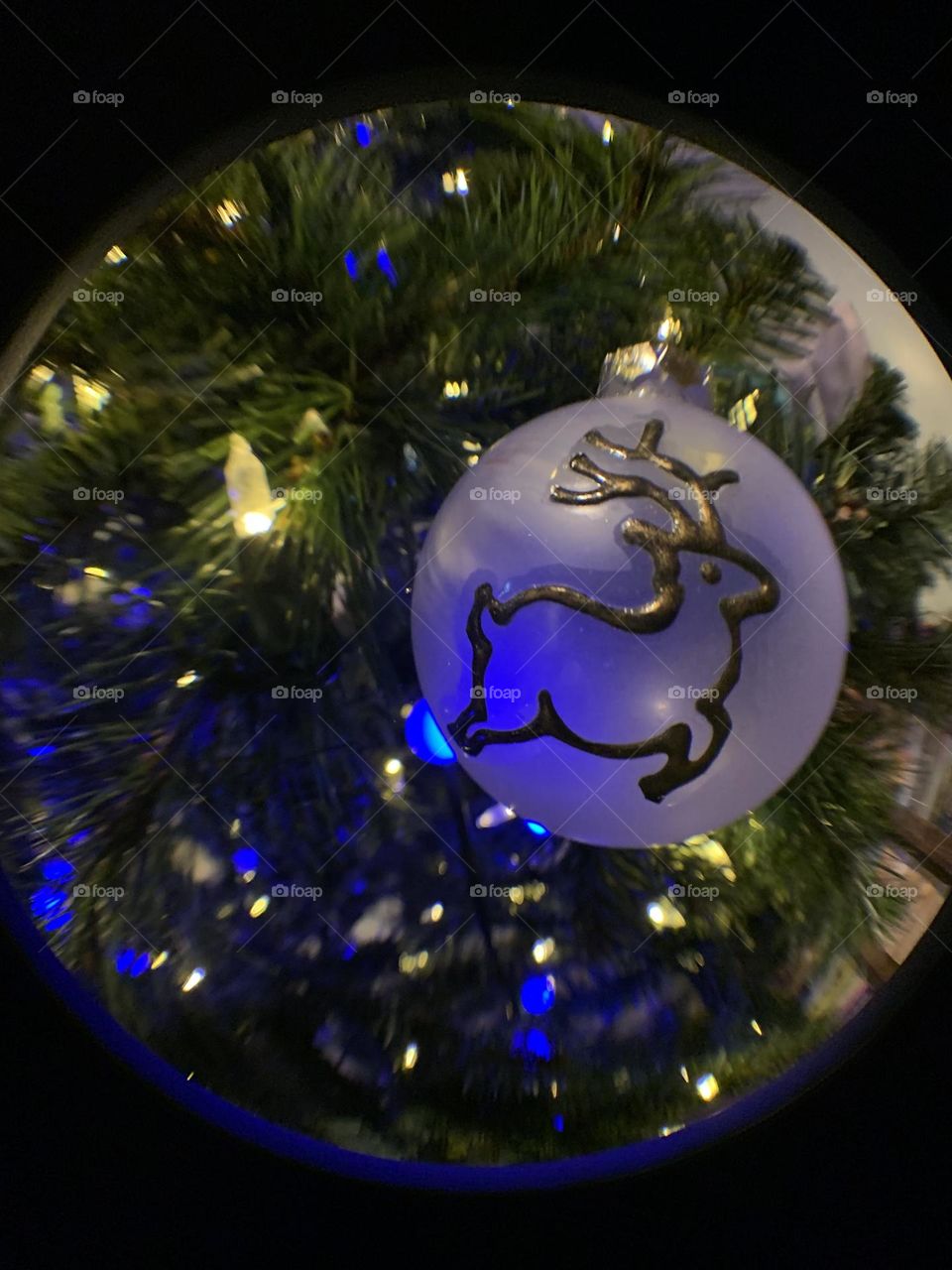 Fisheye lens. Zoom in to a handmade glass ornament, with a Christmas tree behind it. Pretty blue and white lights. 