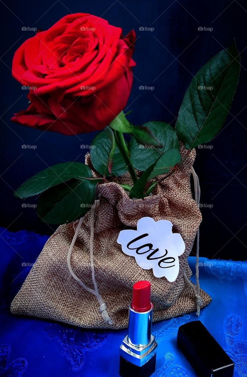 Red lipstick in a tube stands on a blue fabric. Against the background of lipstick is a pouch of burlap, in which there is a red rose with green leaves. On the pouch is a white paper cloud with the inscription: "love". The predominance of red