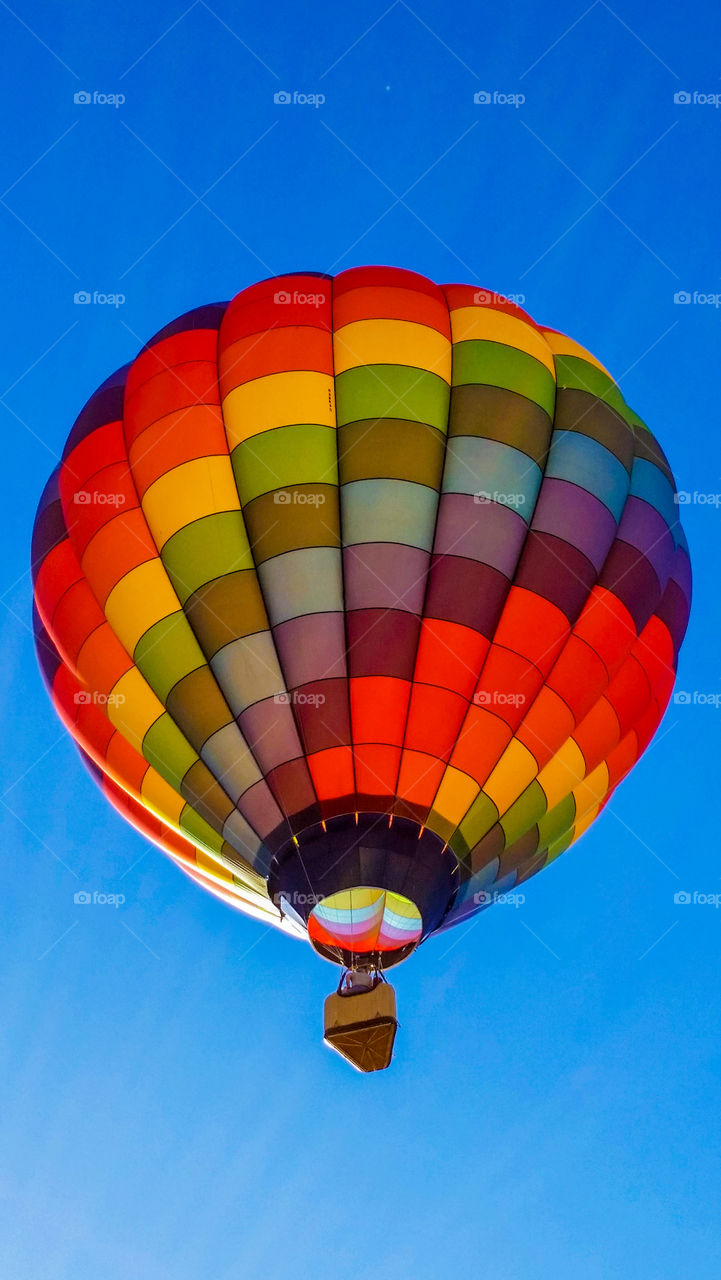 A beautiful rainbow colored hot air balloon ascends into the clear blue sky.