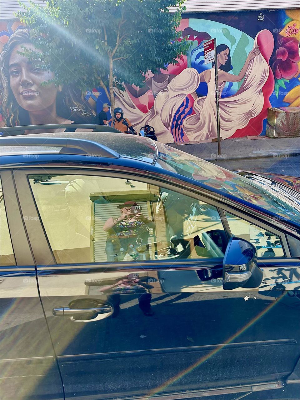 Reflections on the sidewalk of “Jefferson St” off “B’way” in “Bushwick”, Bklyn show me snapping the photo in a mirrored car window and the art mural to Latin “PRIDE” across the street reflects in the metallic blue car roof. 2024. Hypnotic Productions